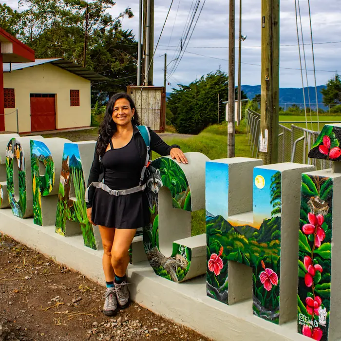 "Maru, guía de Caminantes Acseri, posando junto al rótulo decorado que da la bienvenida a Copalchí."